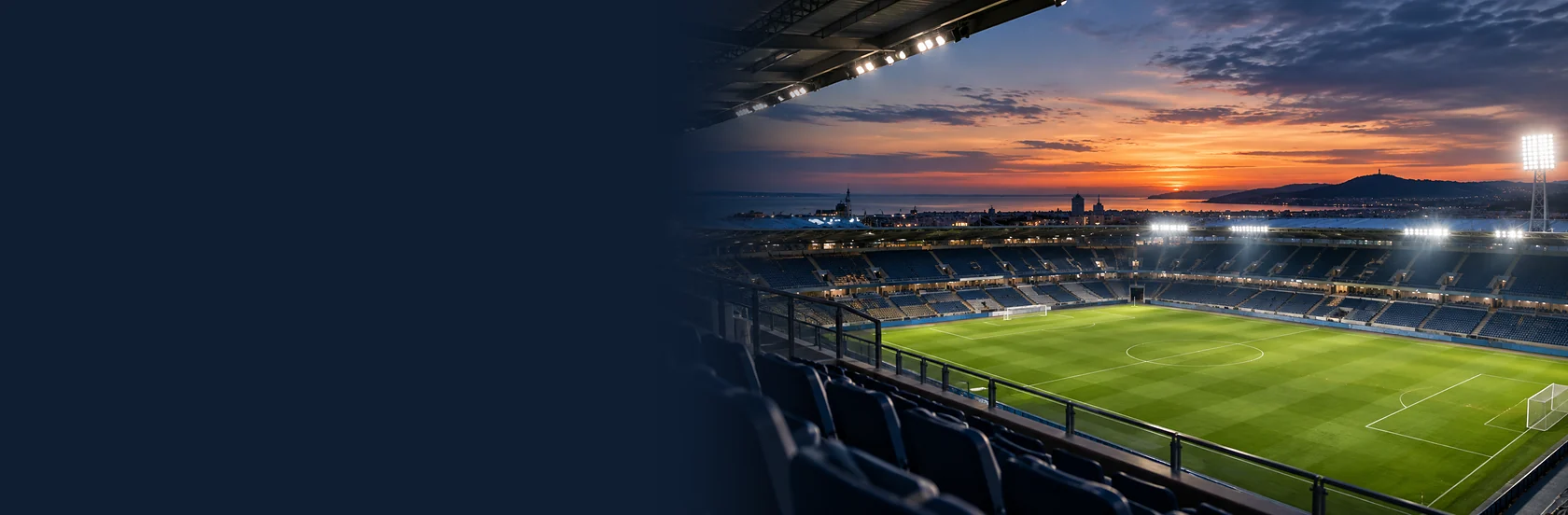A floodlit stadium at sunset seen from the stands, with the coast beyond